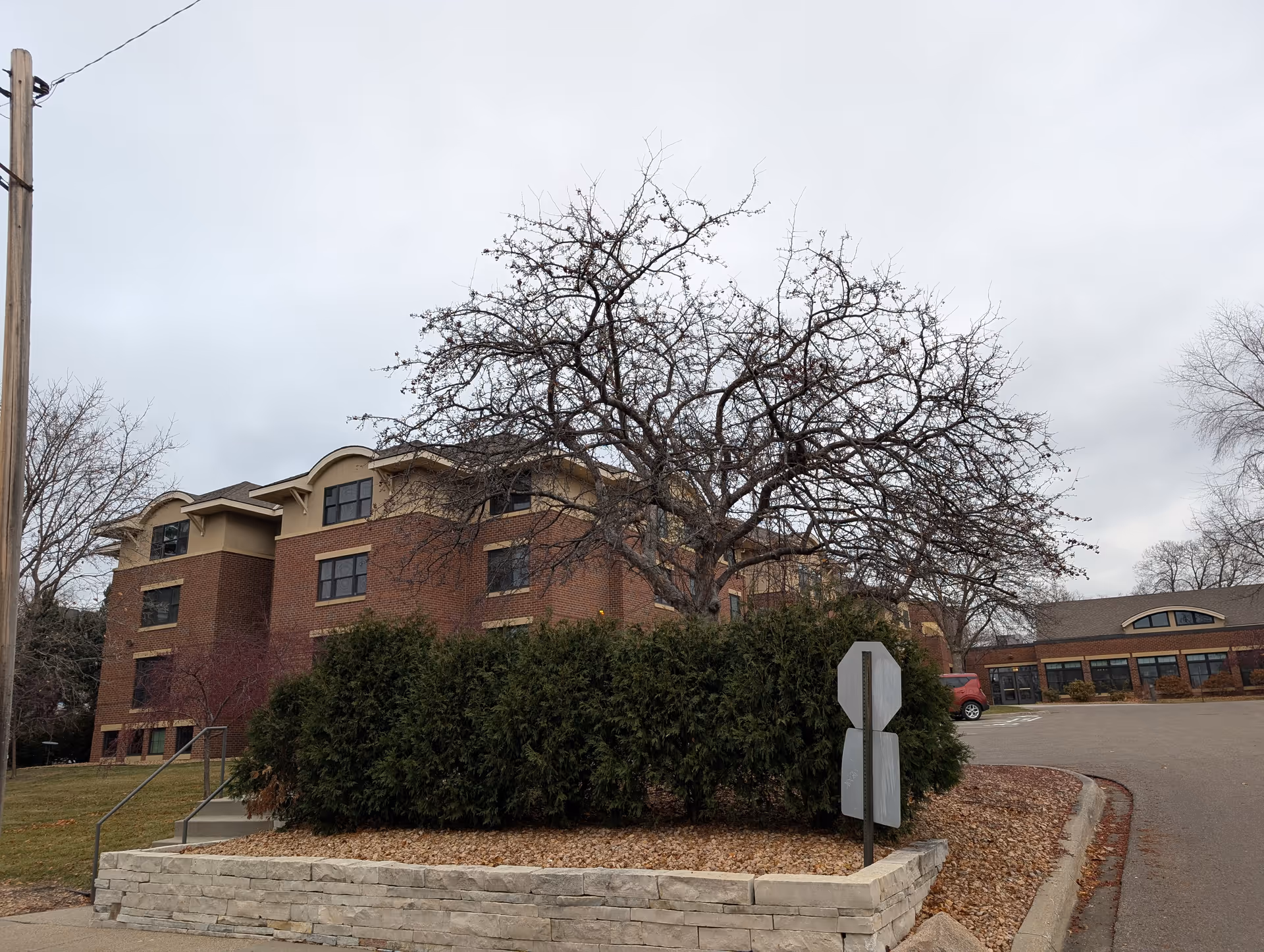 Exterior view of a multi-story brick building with beige trim, surrounded by leafless trees and green bushes. A stone retaining wall and a curved driveway are visible in the foreground under a cloudy sky.