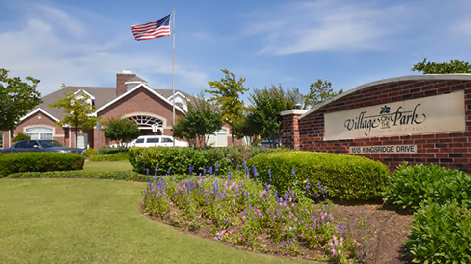 Brick entrance sign and landscaped lawn in front of the Village on the Park senior living building with an American flag flying.