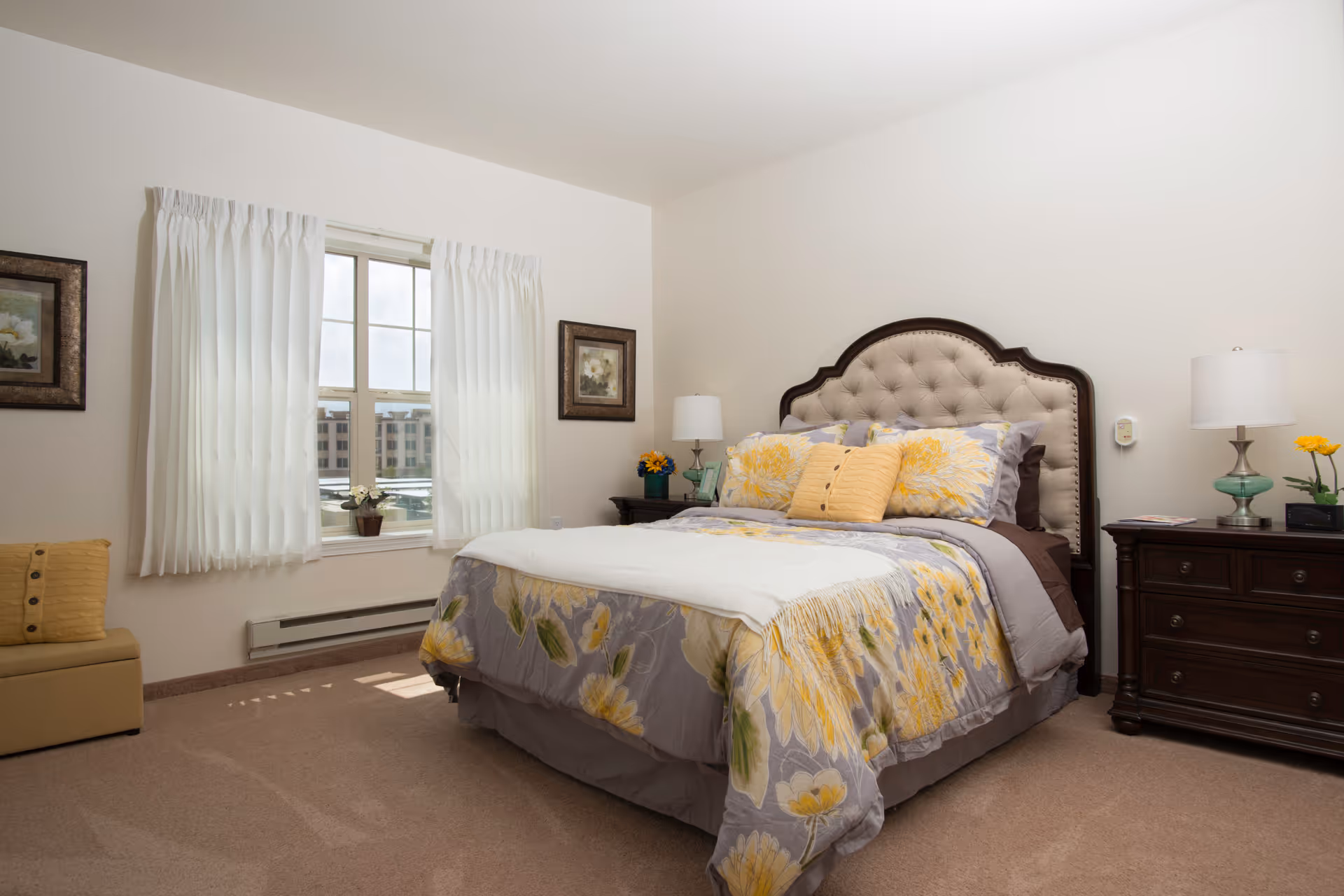 A bright bedroom with a large bed featuring a tufted headboard and floral bedding in shades of yellow and gray. The room has beige carpeting, two dark wooden nightstands with lamps and small flower arrangements, a window with white curtains, and framed artwork on the walls.