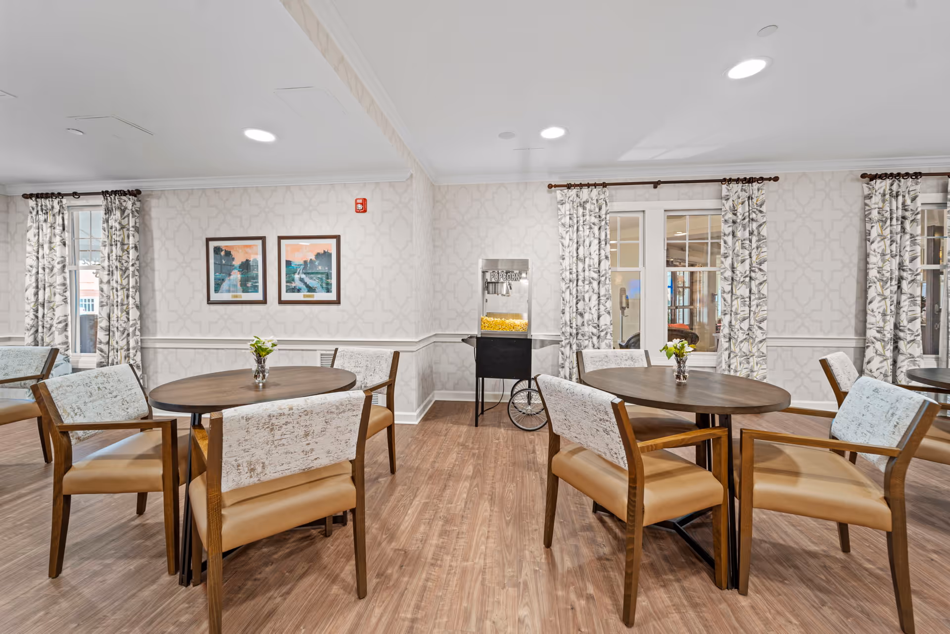 A bright dining room with wooden round tables and cushioned chairs. The room has patterned wallpaper, floral curtains on windows, and a popcorn machine in the corner. Two framed pictures hang on the wall above a chair rail molding.