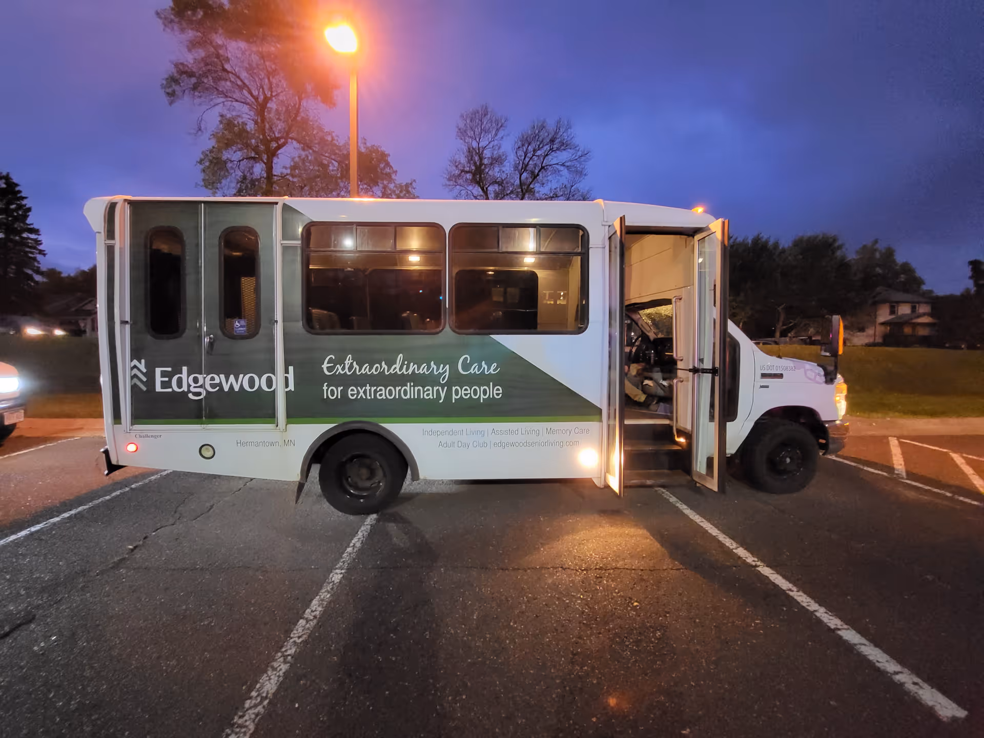 A white and green shuttle bus parked in a parking lot at dusk with its front door open. The bus has the Edgewood logo and the text 'Extraordinary Care for extraordinary people' on its side. There are trees and a streetlamp in the background.