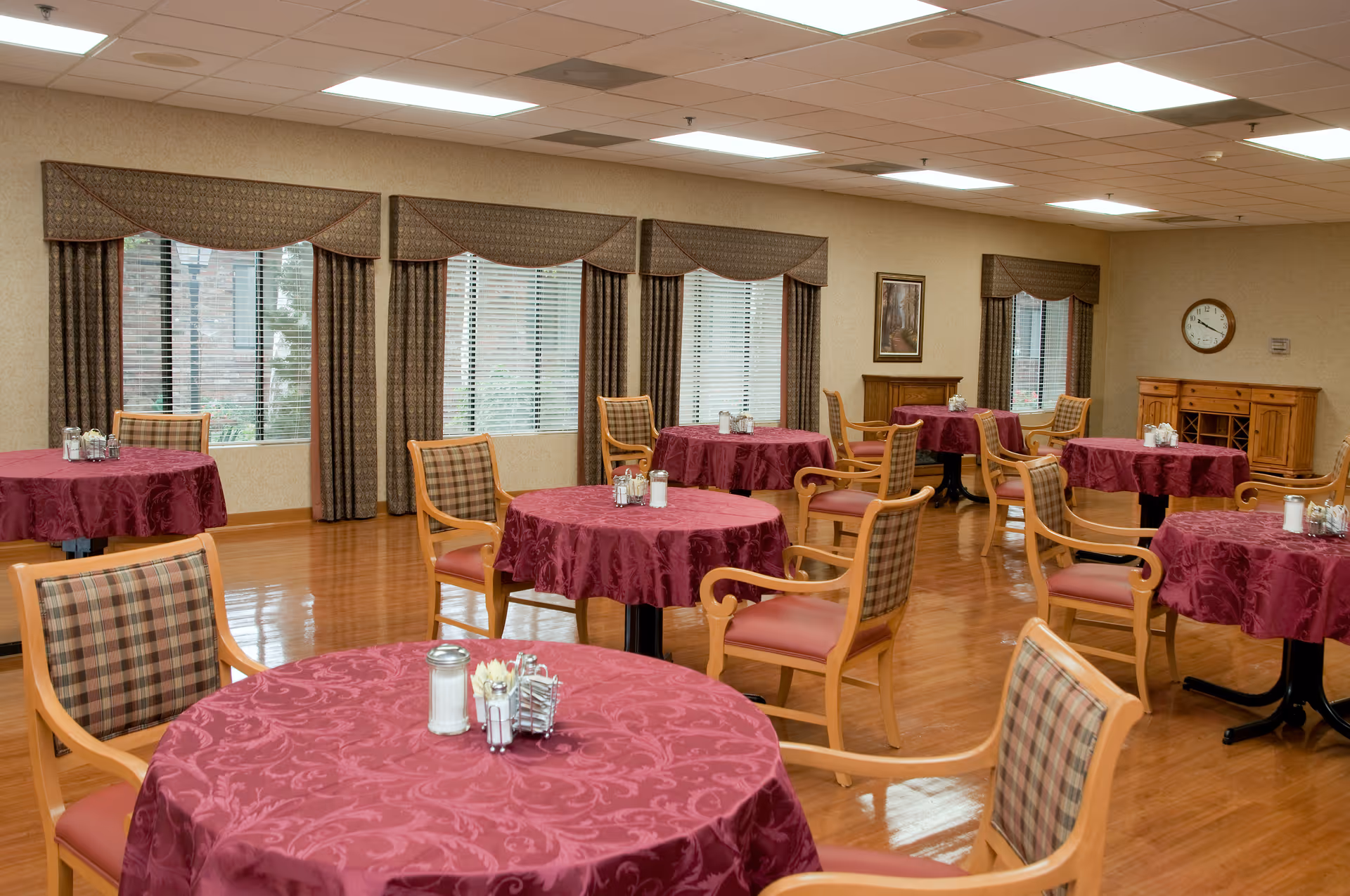 Dining room with round tables covered in burgundy tablecloths and wooden chairs under ceiling lights.