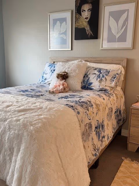 A cozy bedroom with a bed featuring floral patterned bedding in blue and white tones, a fluffy white blanket, and a decorative doll placed on the bed. Above the bed are three framed artworks, two with leaf designs and one with a portrait of a woman. A wooden nightstand is visible to the right side of the bed.