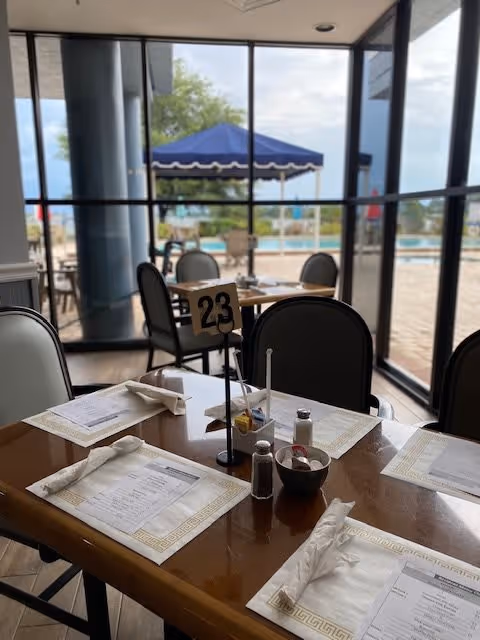 A dining table set for four with menus, napkins, salt and pepper shakers, and a table number 23 in a room with large glass windows showing an outdoor pool area with a blue canopy and patio furniture.