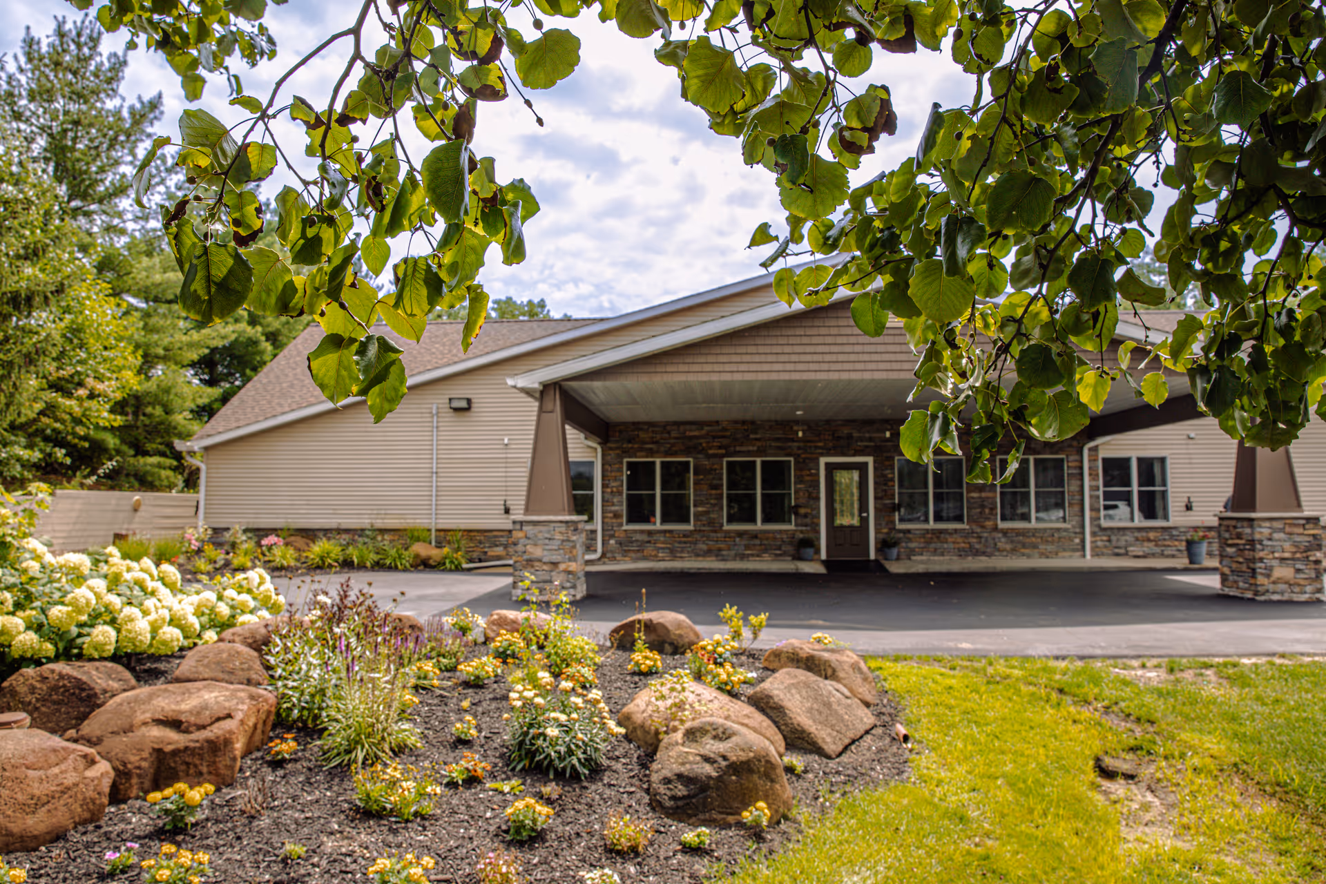 Exterior view of Courtyard Manor of Fenton showing the front entrance of a single-story building with stone and beige siding. The foreground features a landscaped garden with rocks, flowers, and greenery, framed by tree branches with green leaves.