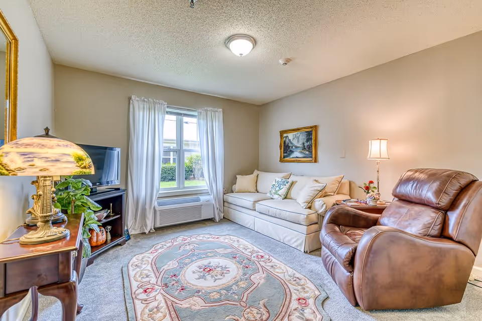 A cozy living room with a beige sofa adorned with white and floral pillows, a brown leather recliner chair, a decorative area rug with floral patterns, a wooden side table with a lamp, a small TV on a stand, and a window with white curtains letting in natural light.