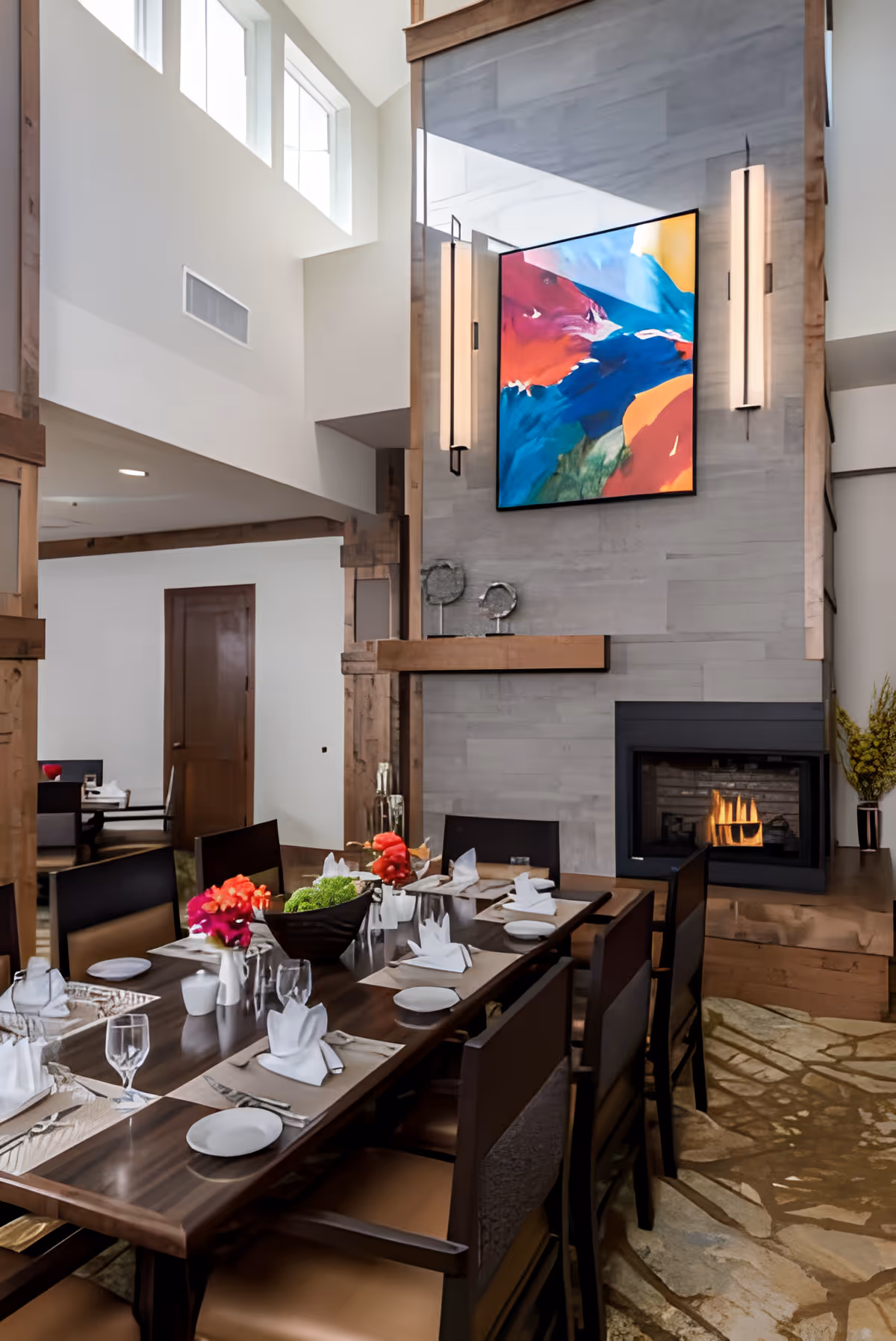 A modern dining room with a long wooden table set for a meal, including plates, glasses, napkins, and cutlery. The room features a tall stone fireplace with a fire burning and a colorful abstract painting above it. The space has high ceilings with large windows allowing natural light to fill the room. There are decorative flowers on the table and contemporary wall sconces on either side of the painting.
