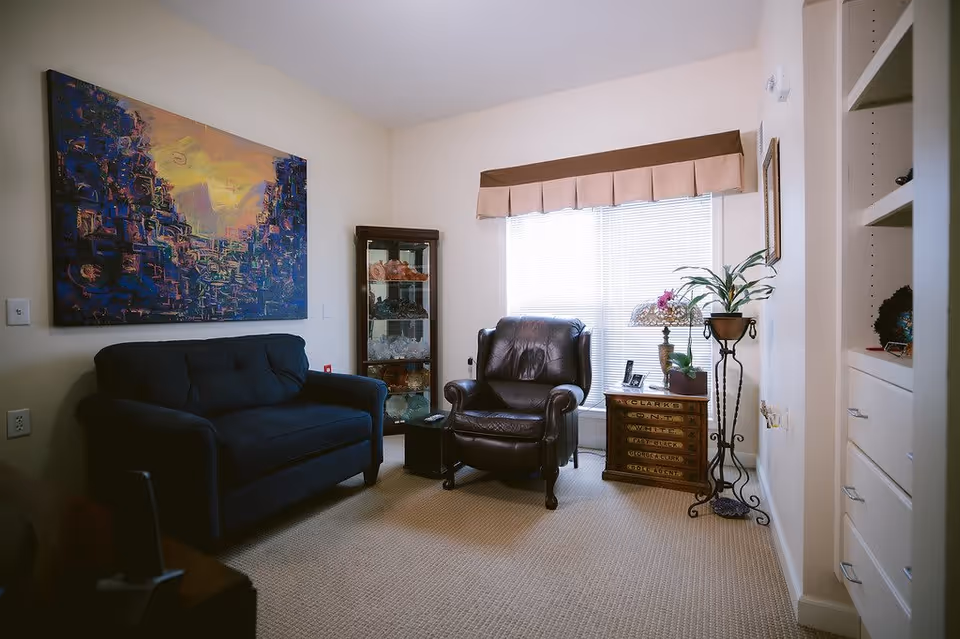 A cozy living room with a dark blue couch on the left and a dark brown leather armchair near a window with blinds and a beige valance. There is a small wooden side table with drawers next to the armchair, holding a lamp and a potted plant. A tall glass cabinet with decorative items is positioned between the couch and the window. The walls are light-colored, and a large abstract painting with dark blue and yellow tones hangs above the couch. The floor is carpeted in a neutral color.