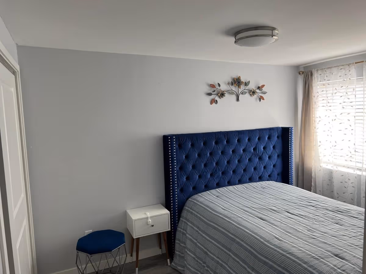 A bedroom with a large bed featuring a tufted blue headboard, a small white nightstand with a drawer, a blue cushioned stool with a metal frame, a window with sheer white curtains, and a decorative metal wall art above the bed.