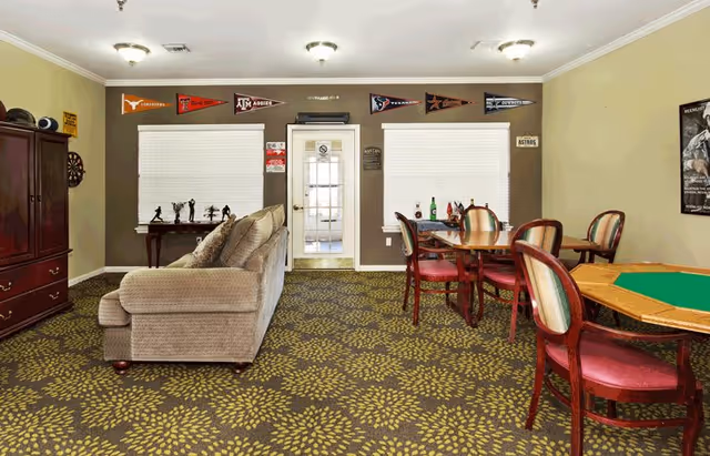 A common area in a senior living facility featuring a beige sofa, a wooden cabinet, a dartboard on the wall, and two tables with chairs. The walls are decorated with sports pennants and a poster. The carpet has a floral pattern, and there is a door with windows in the center of the back wall.