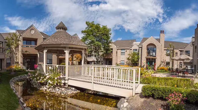 Outdoor view of a senior living facility with a wooden gazebo connected by a white railing bridge over a small pond. The background shows a multi-story brick building with balconies, windows, and landscaped gardens under a partly cloudy blue sky.