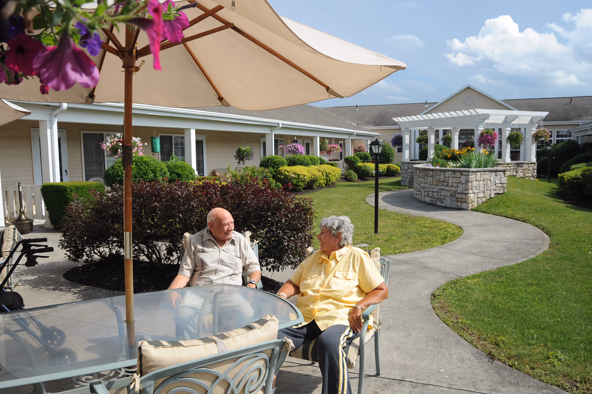 Two elderly people sitting and talking at a glass outdoor table under a large beige umbrella in a well-maintained garden courtyard with a curved concrete pathway, green bushes, hanging flower baskets, and a white pergola structure in the background.