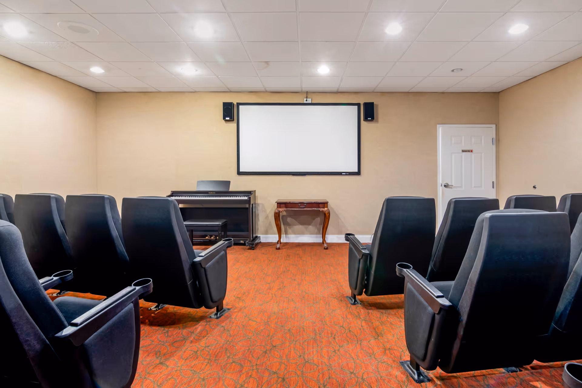 A small theater room with rows of black cushioned seats facing a blank projector screen mounted on a beige wall. There is a digital piano and a small wooden table beneath the screen. The room has a red patterned carpet and a white door on the right side.
