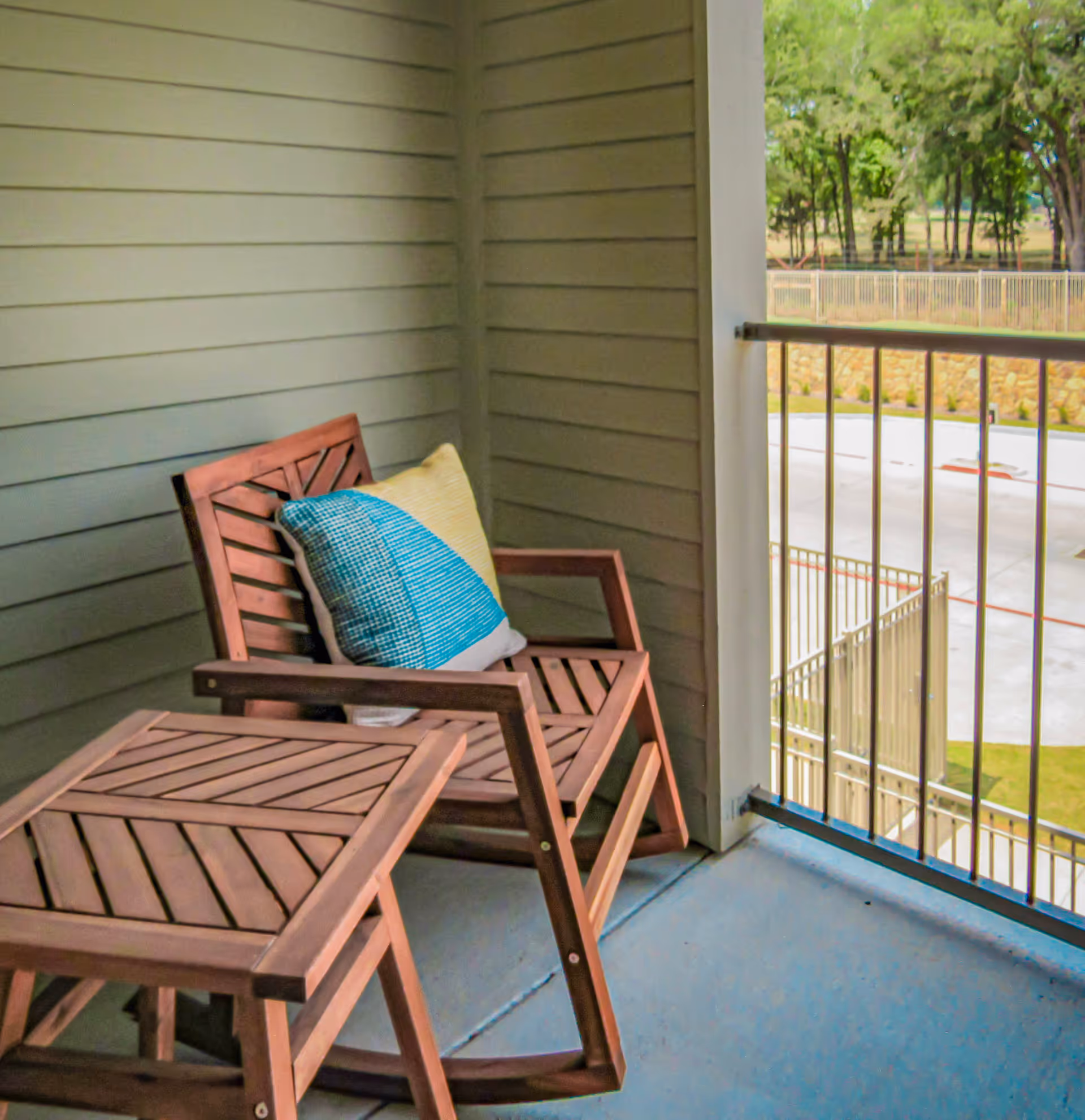 A small outdoor balcony area with a wooden rocking chair featuring two cushions, one blue and one yellow, and a matching wooden table. The balcony has a metal railing and overlooks a parking area and trees in the background.