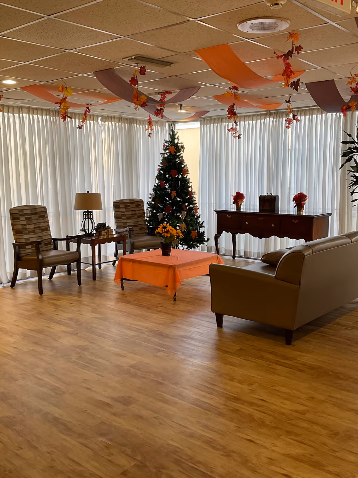 A cozy living room area with wooden flooring, featuring a decorated Christmas tree in the corner. There are two patterned armchairs with a small table and lamp between them, a wooden console table with two flower vases, and a beige sofa. An orange tablecloth covers a small coffee table with a flower pot on it. The ceiling is decorated with orange and brown streamers and hanging autumn leaves. Large windows with white curtains allow natural light to fill the room.