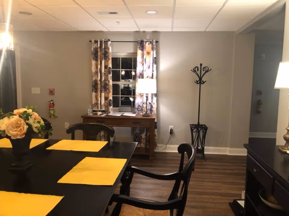 A dining area featuring a black table with yellow placemats and a floral centerpiece, chairs, a desk beneath a curtained window, and a coat rack against a gray wall.
