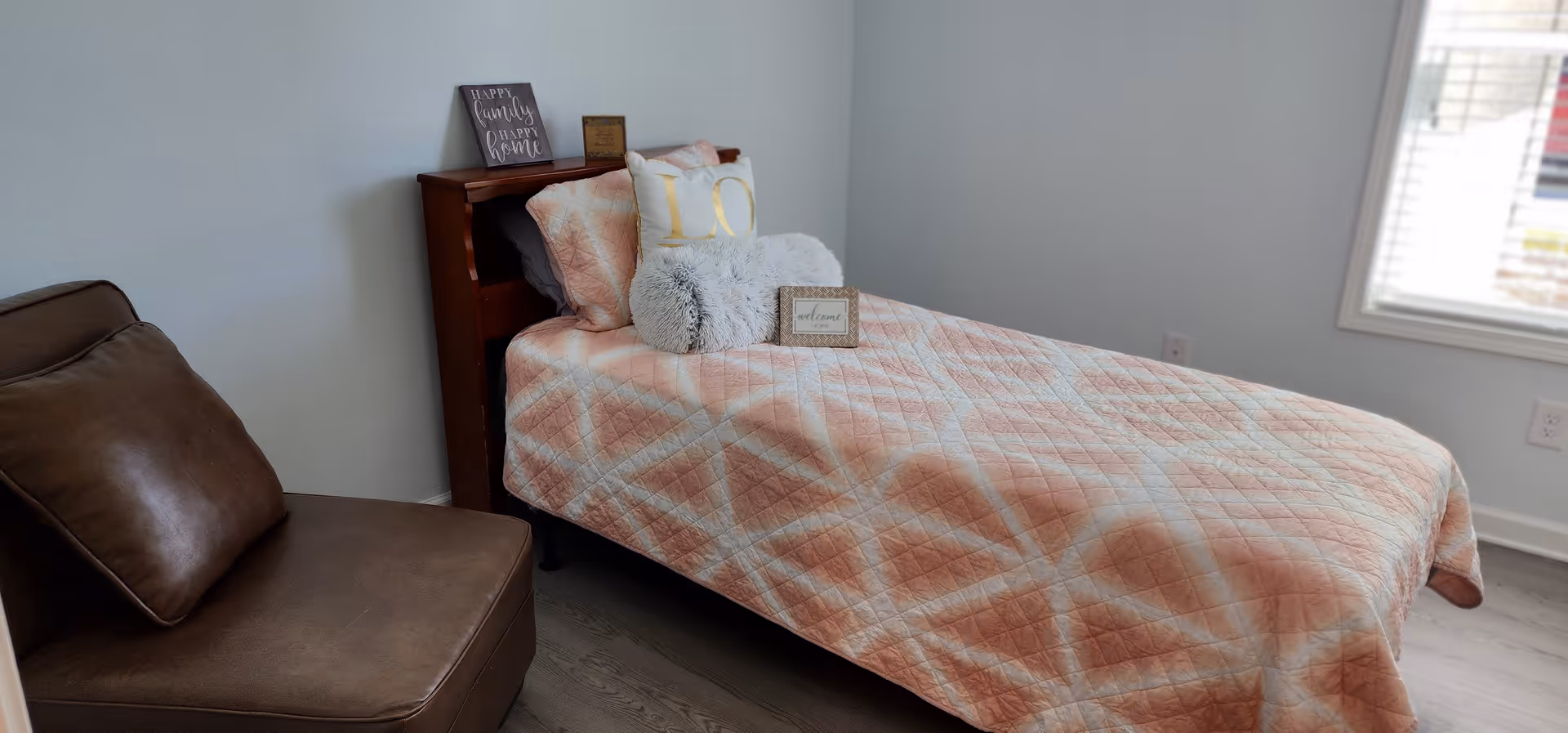 A neatly made single bed with a peach and white geometric patterned quilt, several decorative pillows including a fluffy gray one and a white one with gold letters, and a small framed welcome sign placed on the bed. Next to the bed is a brown leather chair. The room has light-colored walls and a window with blinds letting in natural light.