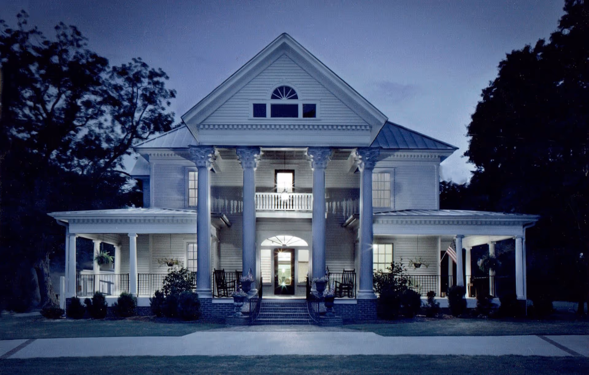 Front exterior view of a large, white, two-story house with classical columns and a porch with rocking chairs, illuminated at dusk with trees on either side.