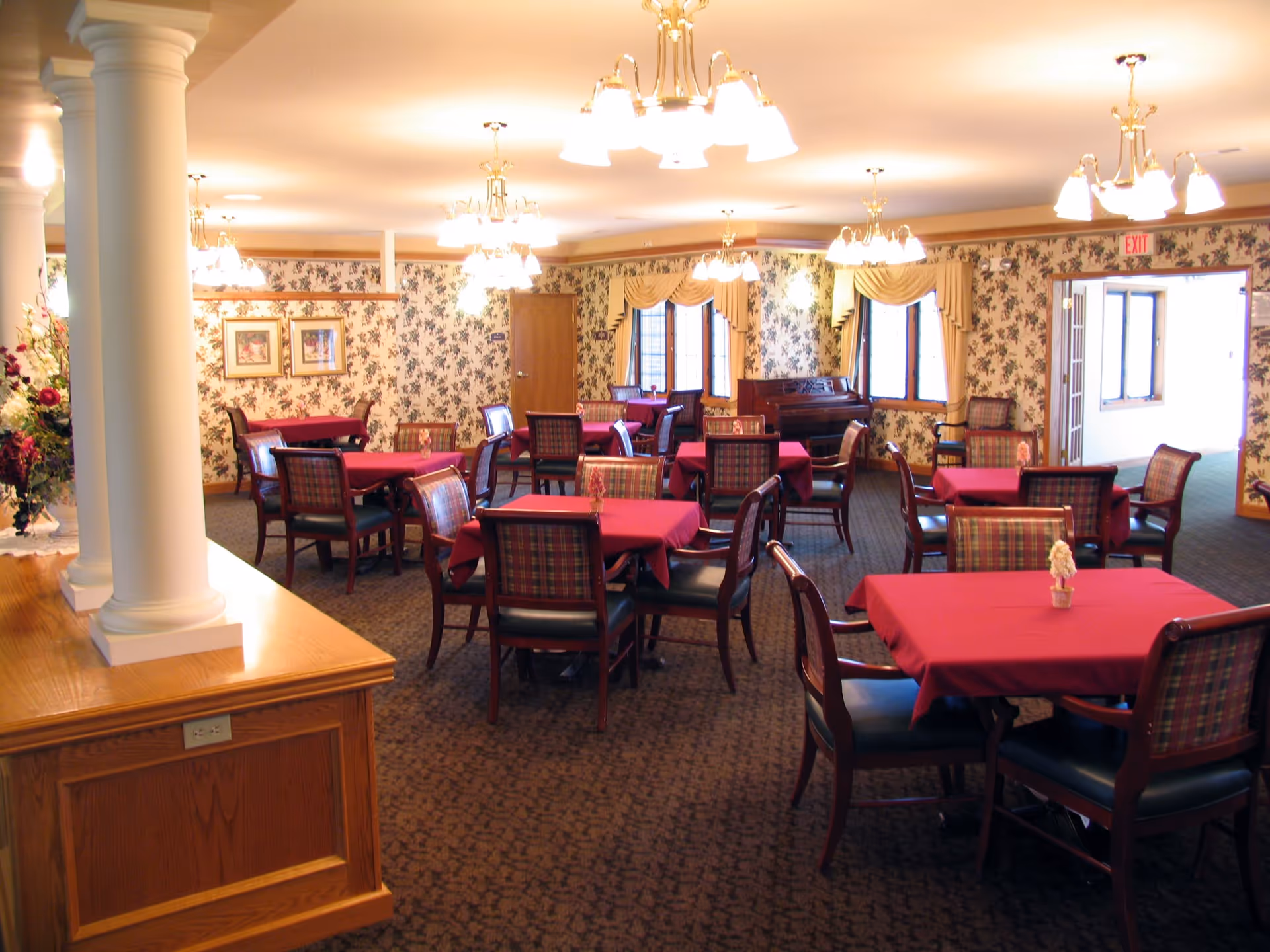A dining room with several tables covered in red tablecloths, each surrounded by wooden chairs with plaid upholstery. The room features floral wallpaper, multiple chandeliers, large windows with draped curtains, and a piano in the corner. There are white columns and a wooden counter on the left side of the image.