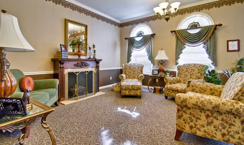 A cozy senior living facility lounge with floral upholstered armchairs, a green sofa, a wooden fireplace with a decorative screen, and two arched windows with green drapes. The room is carpeted with a patterned design and has warm beige walls with a decorative border near the ceiling.