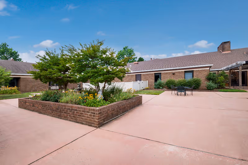 Brick courtyard with a raised planter bed, patio table and chairs, and surrounding single-story brick buildings under a blue sky.