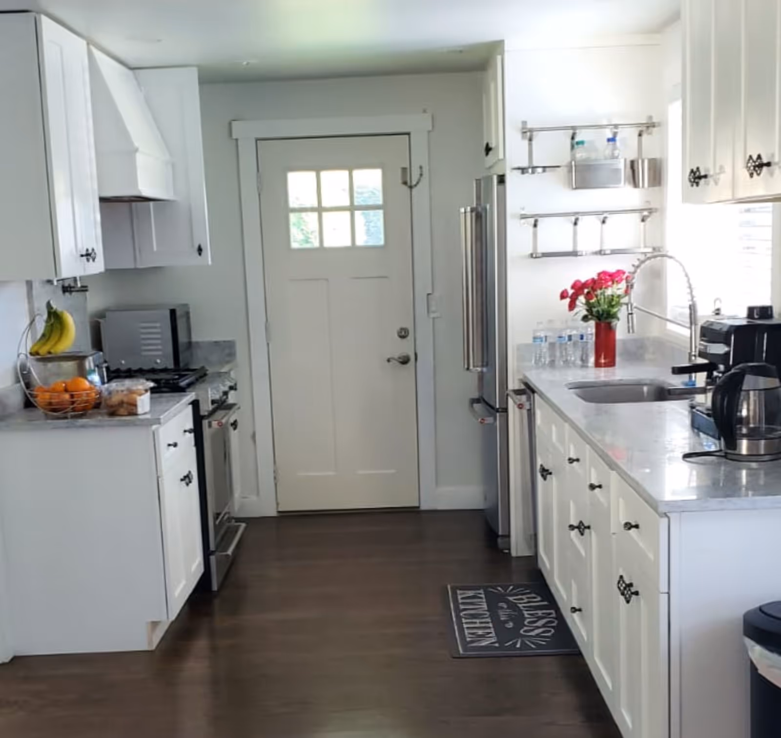 A bright kitchen with white cabinets, a stainless steel refrigerator, a stove, a microwave, and a sink. There are bananas and oranges in a fruit basket on the counter, a red vase with flowers near the sink, and several water bottles. The floor is dark wood, and there is a door with a window at the end of the kitchen.