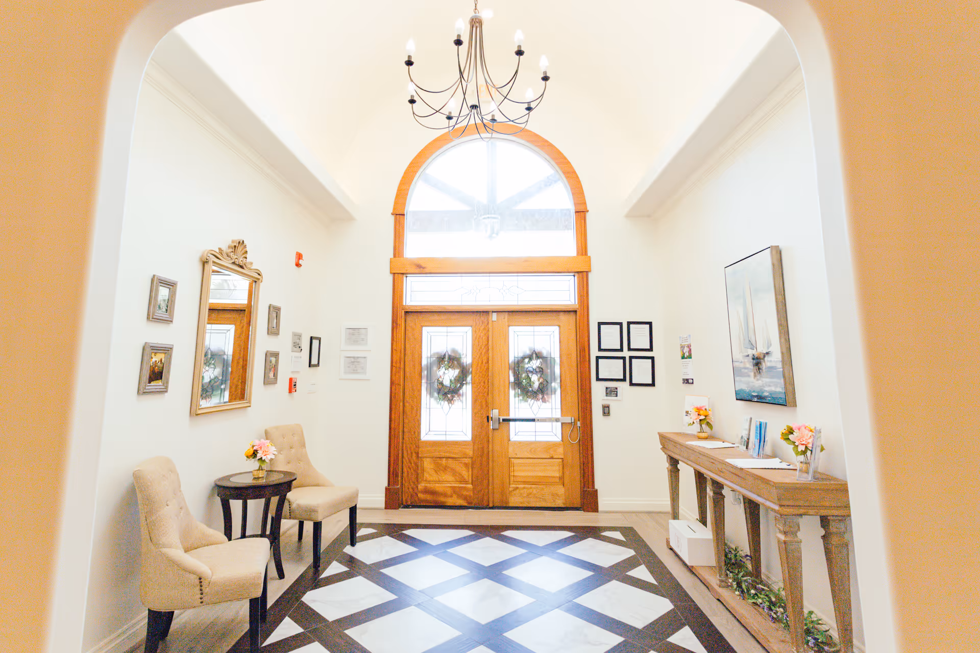 Bright entry foyer with arched double wooden doors decorated with wreaths, a chandelier, seating chairs, and a console table with flowers.