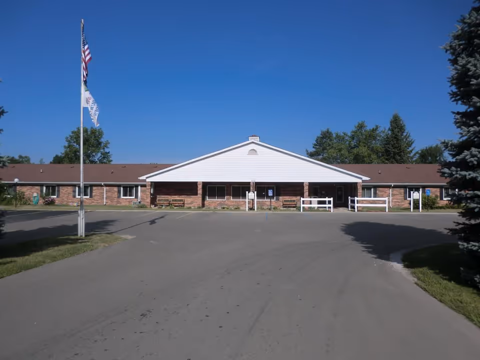 Front exterior view of a single-story brick building with a white triangular roof section above the entrance. There are two flagpoles with flags in front of the building, a paved driveway, and some trees on either side under a clear blue sky.