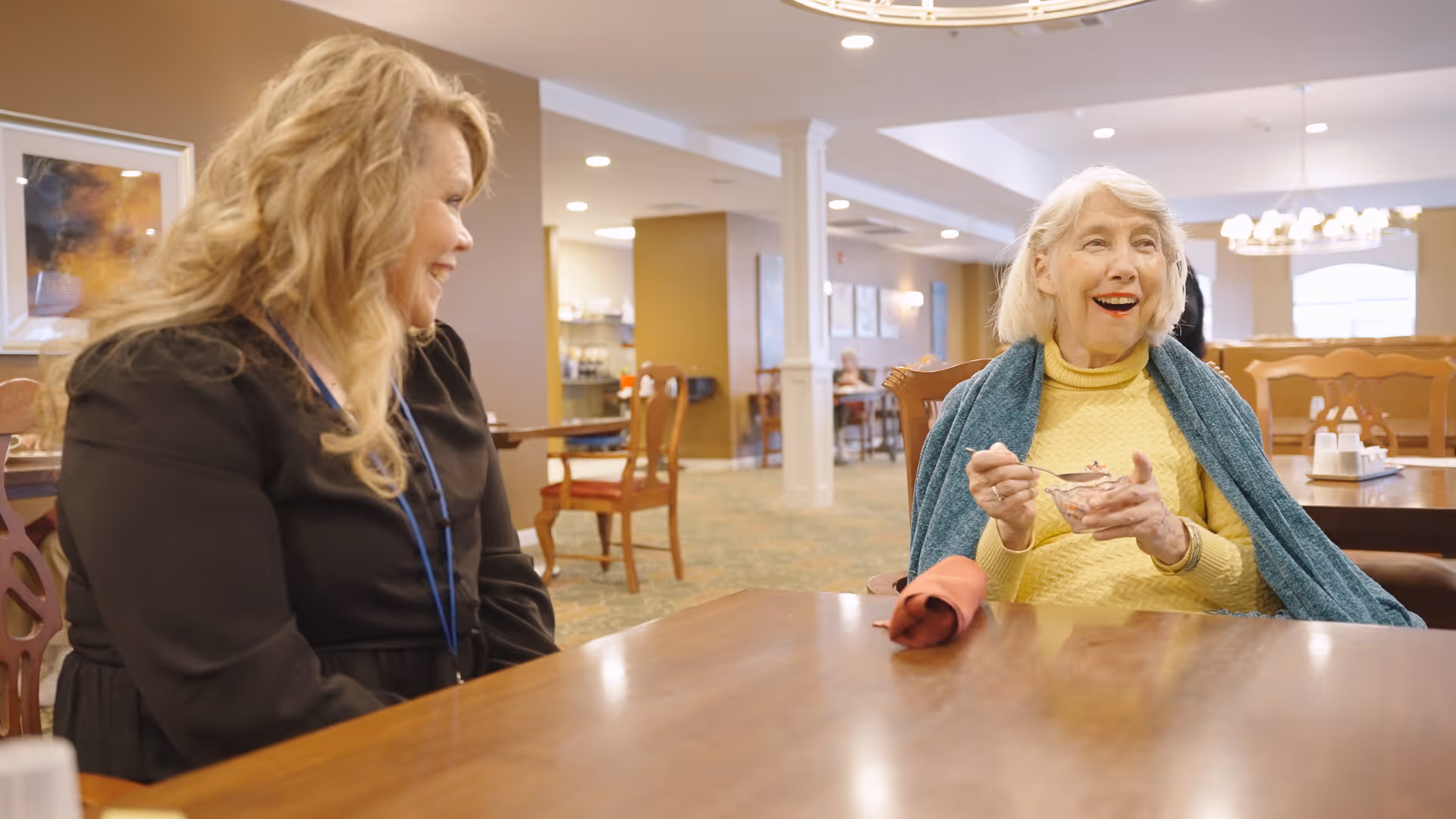 An elderly woman wearing a yellow sweater and teal shawl is sitting at a wooden table eating from a bowl, smiling and engaged in conversation with a woman with long blonde hair wearing a black top and a blue lanyard, in a well-lit dining area with wooden chairs and tables.