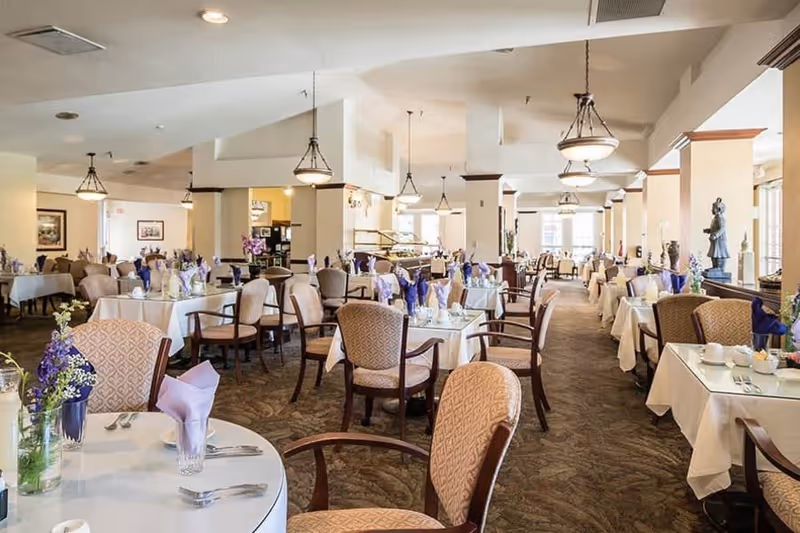 A spacious dining room with multiple tables covered in white tablecloths, each set with purple napkins, glassware, and silverware. The room features cushioned chairs with wooden arms, carpeted floors, hanging light fixtures, and large windows allowing natural light to fill the space. Decorative statues and framed artwork are visible along the walls.