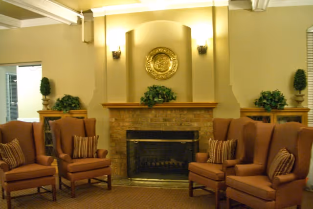 A cozy living room area with a brick fireplace in the center, flanked by two wooden cabinets with green plants on top. Four brown upholstered armchairs with striped cushions are arranged around the fireplace. The walls are painted beige and decorated with a round golden emblem above the fireplace.