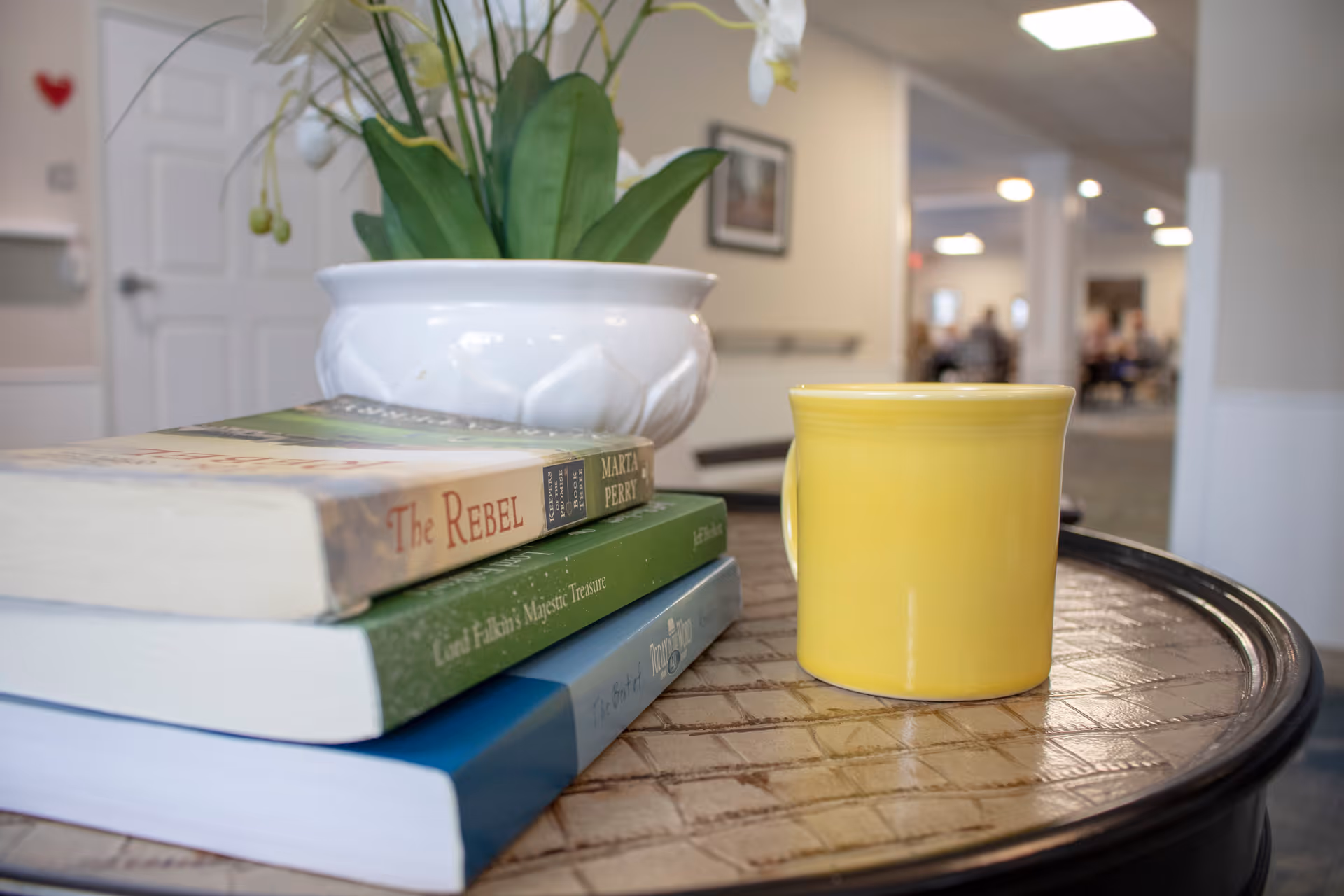 Close-up of a round table with a yellow coffee mug, a stack of three books, and a white ceramic pot with green plants. In the background, there is a hallway with people sitting and talking in a common area.