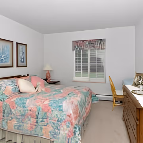 A bedroom with a bed covered in a floral patterned bedspread in shades of pink, green, and blue. There are two framed pictures on the wall above the bed, a bedside table with a lamp, a window with a valance, and a wooden dresser with a chair beside it.