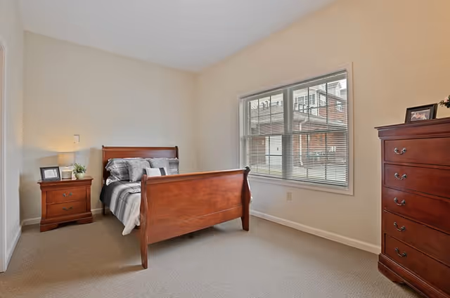 Bright bedroom with a wooden sleigh bed, matching nightstand and dresser, and a window with blinds.
