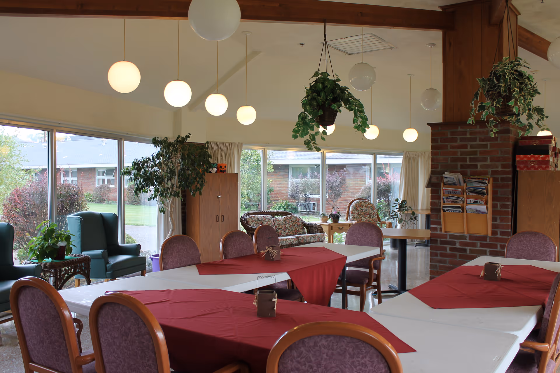 A bright and spacious common area in a senior living facility with large windows showing an outdoor garden. The room features several tables covered with white tablecloths and red runners, surrounded by purple cushioned chairs. There are hanging round light fixtures and green plants, including a hanging plant and potted plants. Comfortable armchairs and a floral-patterned sofa are visible near the windows, along with a brick column holding a magazine rack.