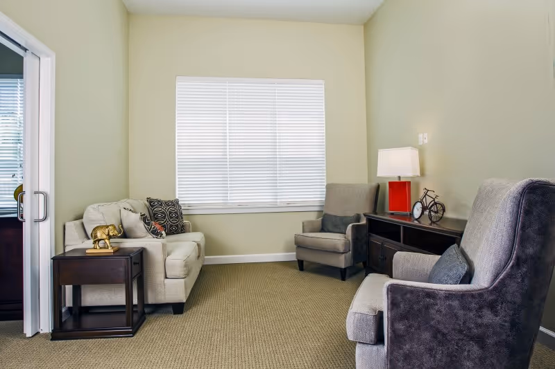 A cozy living room with beige walls and carpeted floor, featuring a beige loveseat with decorative pillows, two gray armchairs, a dark wooden side table with a golden elephant figurine, and a dark wooden console table with a red lamp, a small bicycle decoration, and a clock. A large window with white blinds is centered on the back wall.