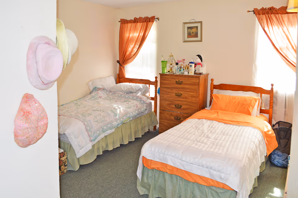 A sunlit bedroom with two twin beds, a wooden dresser between them, orange curtains, and hats hanging on the left wall.