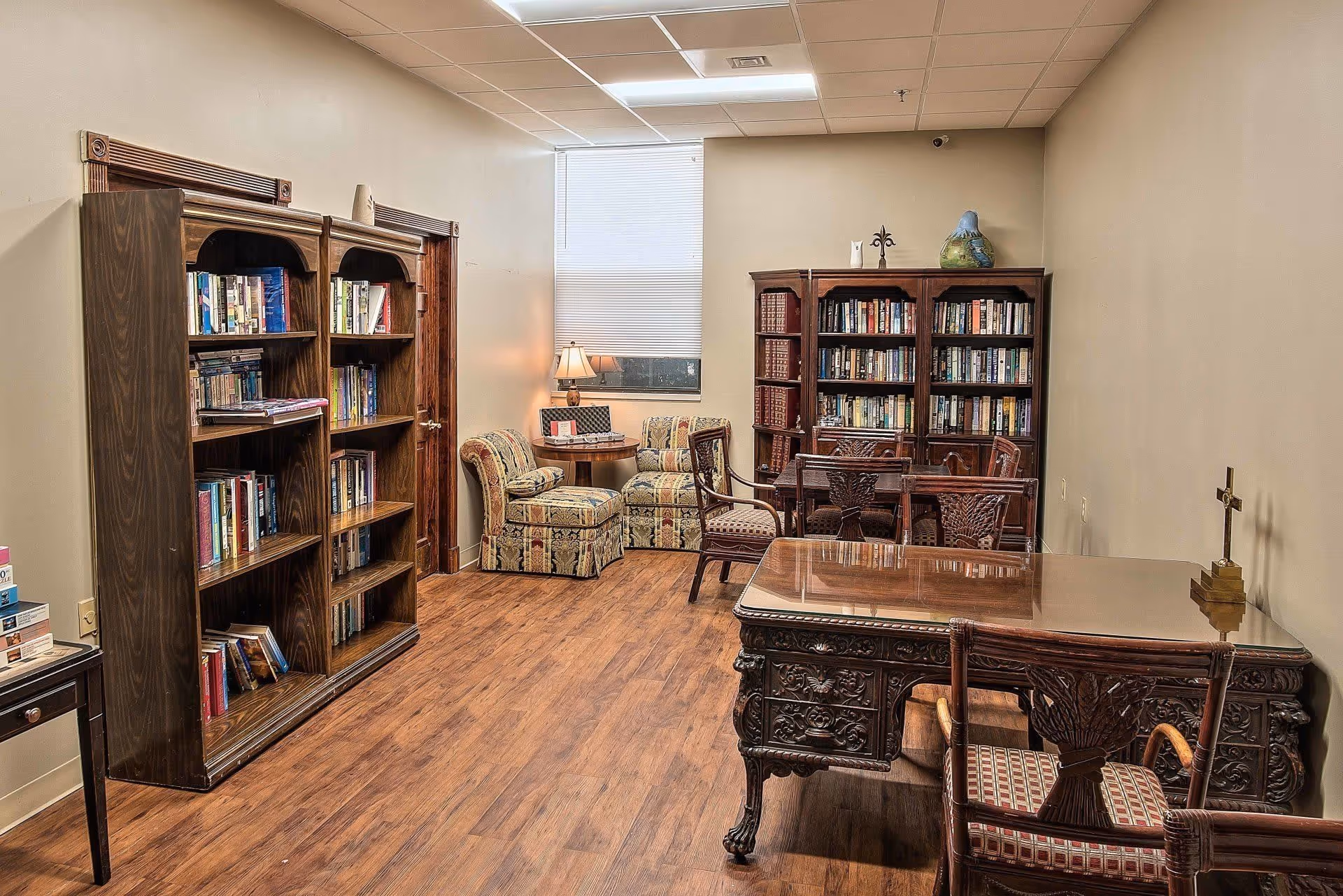 A cozy room with wooden bookshelves filled with books, a wooden desk with ornate carvings, a matching chair, a small table with two upholstered armchairs, a lamp, and a window with a closed blind. The floor is wooden, and the walls are painted beige.