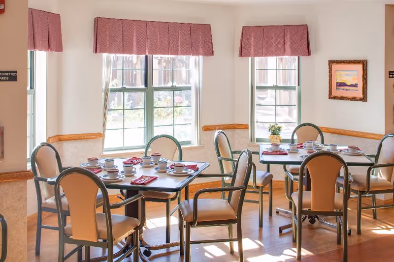 A bright dining room with two tables set for a meal, each with cups, saucers, and red napkins. The room has large windows with red valances letting in natural light, beige walls with wooden trim, and several chairs around the tables. A framed painting and a small potted plant are visible on the walls and tables.