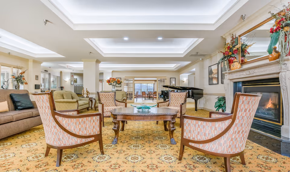 A spacious senior living community lounge with patterned carpet, a set of four upholstered armchairs around a wooden coffee table, a brown sofa, and a green loveseat. There is a fireplace with a decorative mantle and floral arrangements on the right side, and a grand piano in the background. The room has recessed ceiling lights and large windows letting in natural light.