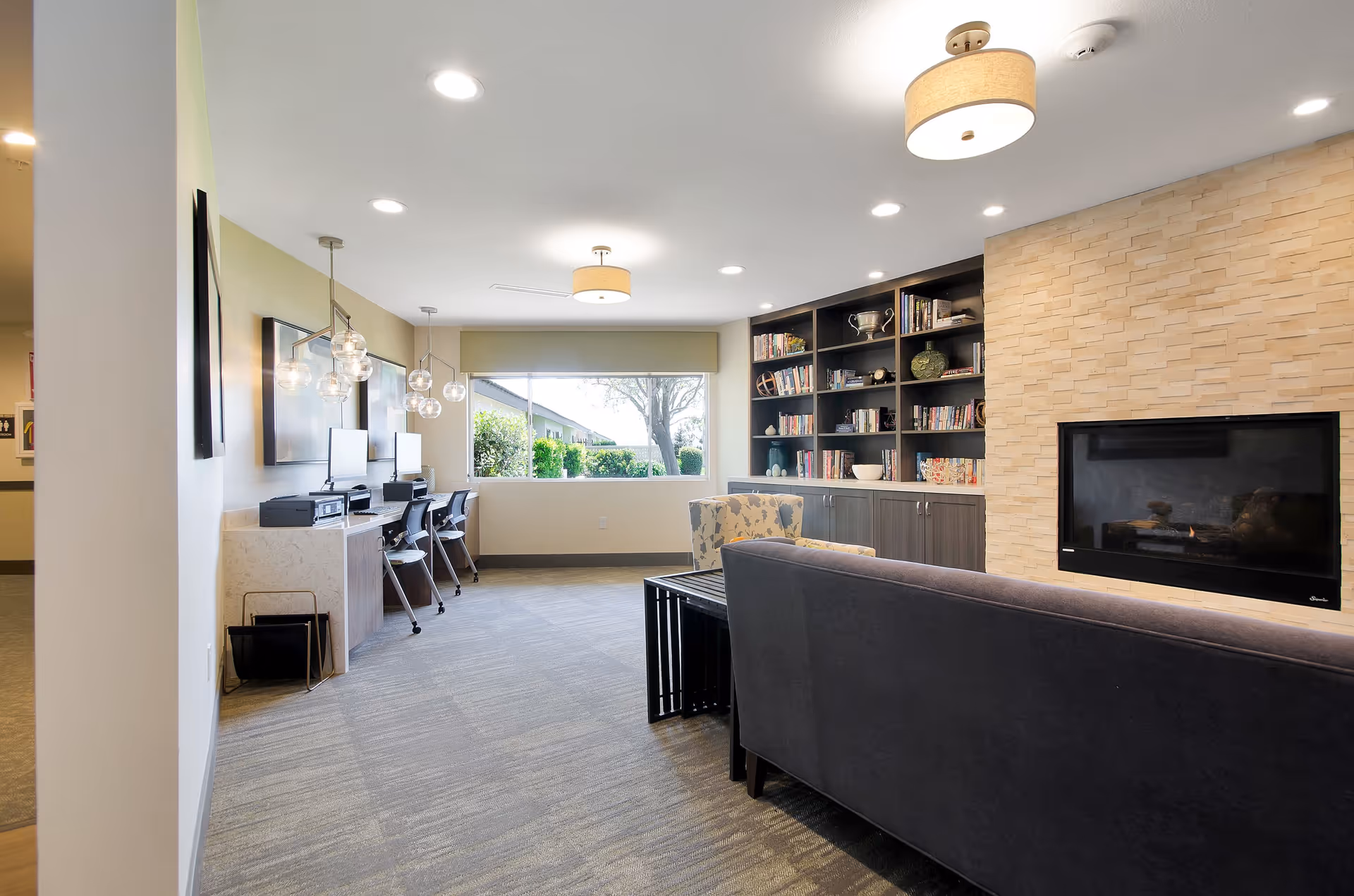 A cozy common area in an assisted living facility featuring a dark gray couch facing a modern fireplace set in a light stone wall. To the right of the fireplace is a built-in bookshelf filled with books and decorative items. On the left side of the room, there are three computer workstations with chairs and pendant lights hanging above. A large window at the back lets in natural light and shows greenery outside. The room has recessed ceiling lights and neutral-colored walls and carpet.