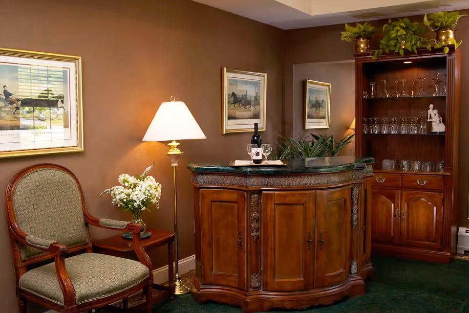A cozy interior corner featuring a vintage wooden bar with a green marble top, a bottle of wine and two glasses on a tray. To the left, there is an upholstered wooden armchair next to a small side table with a vase of white flowers and a tall floor lamp. The walls are painted brown and decorated with framed pictures of horses. On the right, a wooden cabinet with glass shelves holds various glassware and a small white statue, topped with green plants.