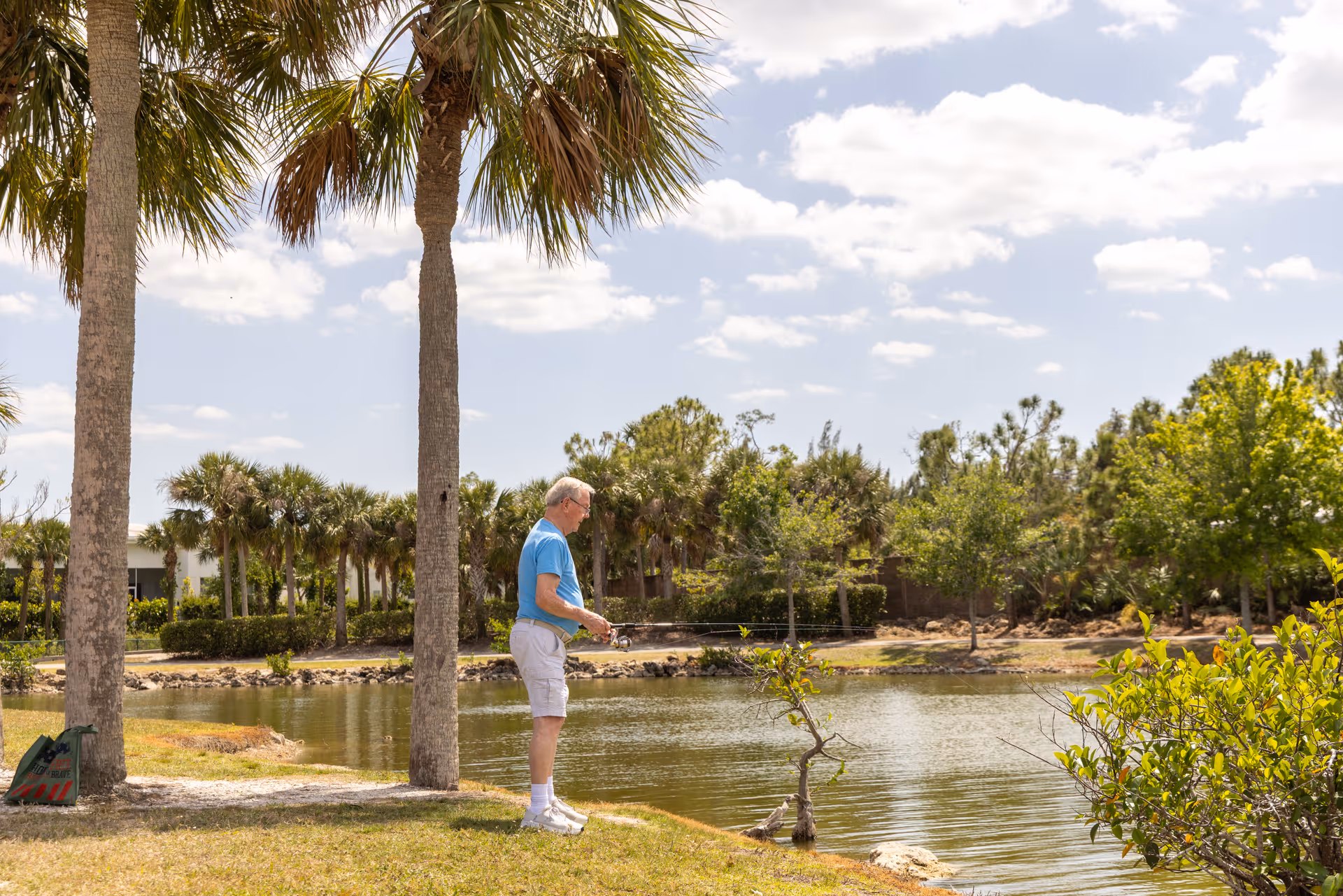 An elderly man wearing a blue shirt and white shorts is fishing by a pond surrounded by palm trees and greenery under a partly cloudy sky.