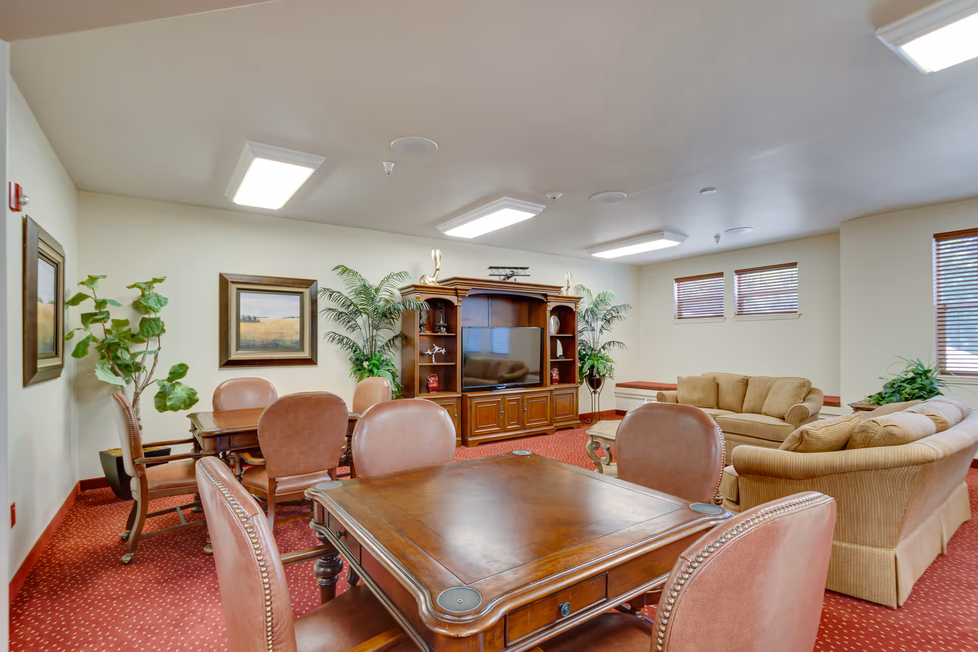A cozy living room area in Magnolia Heights Gracious Retirement Living featuring a wooden game table with four leather chairs in the foreground, a second table with chairs against the wall, a large wooden entertainment center with a flat-screen TV, beige sofas, potted plants, framed artwork, and windows with wooden blinds. The room has a red patterned carpet and bright ceiling lights.