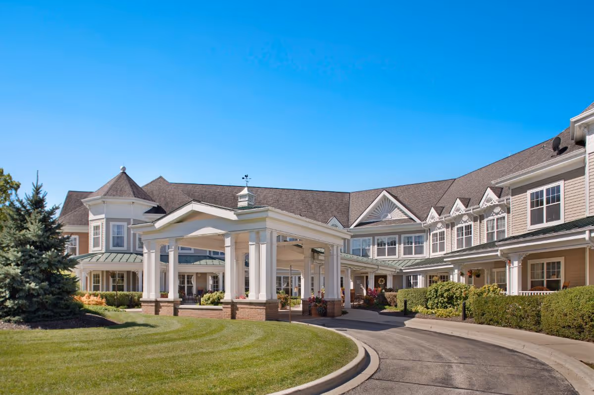 Exterior view of a large senior living facility building with a covered entrance, manicured lawn, shrubs, and clear blue sky.