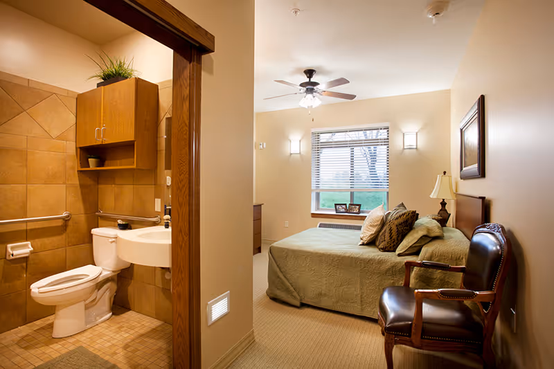 Well-lit bedroom with a bed, armchair, and an accessible bathroom visible through an open doorway.
