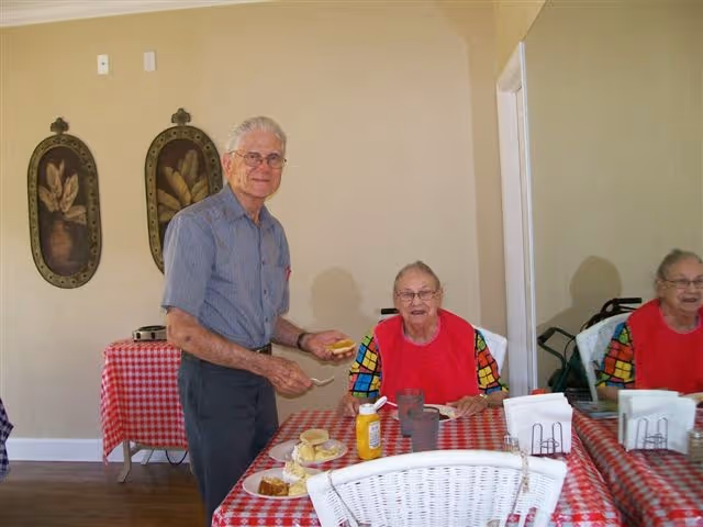 Two elderly residents at a red-checkered dining table with plates, condiments, and a mirror on the wall in a communal dining area.