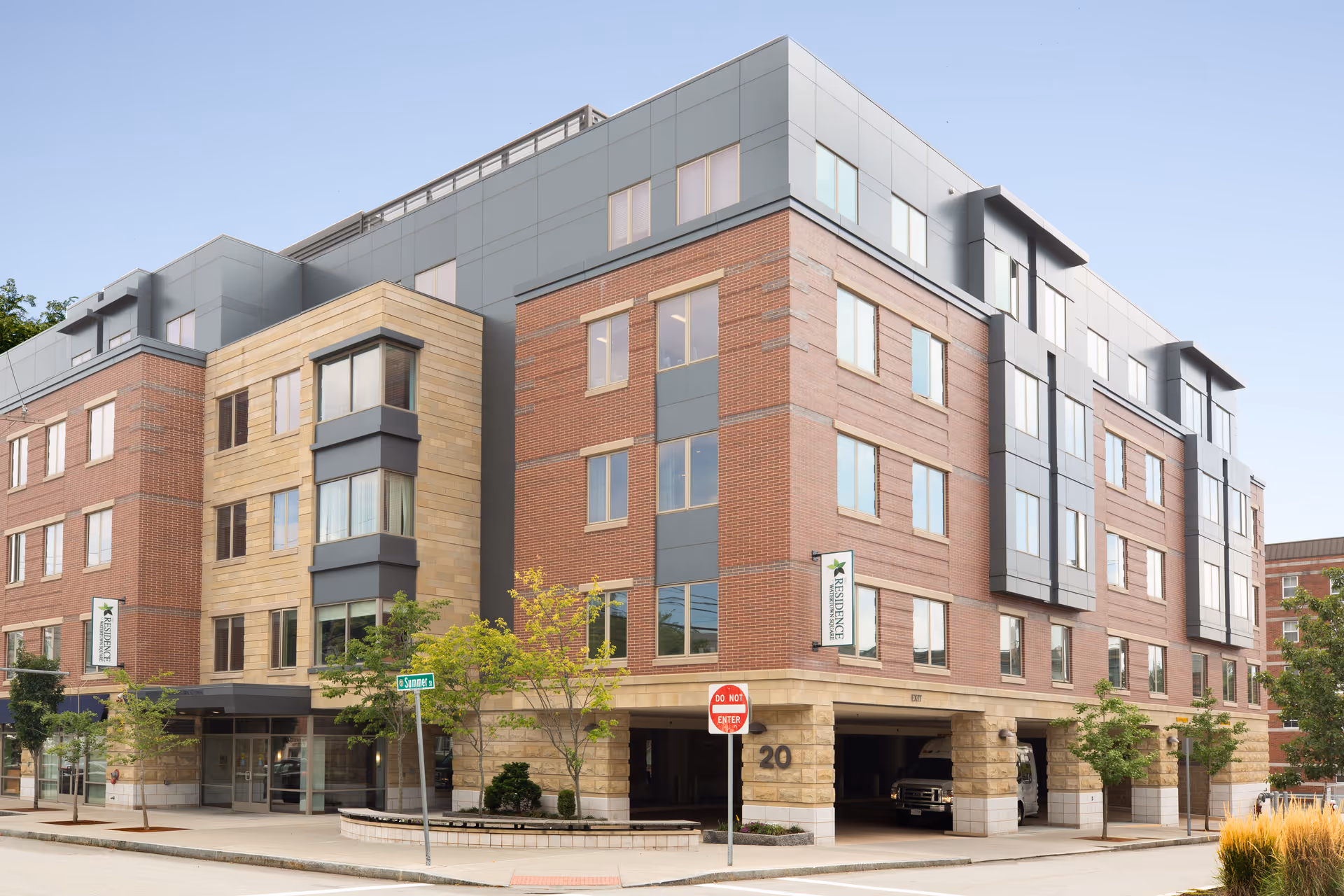 Exterior view of a modern multi-story senior living facility named The Residence at Watertown Square, featuring a combination of brick and stone facade with large windows and a covered parking area on the ground floor. Trees and street signs are visible along the sidewalk.