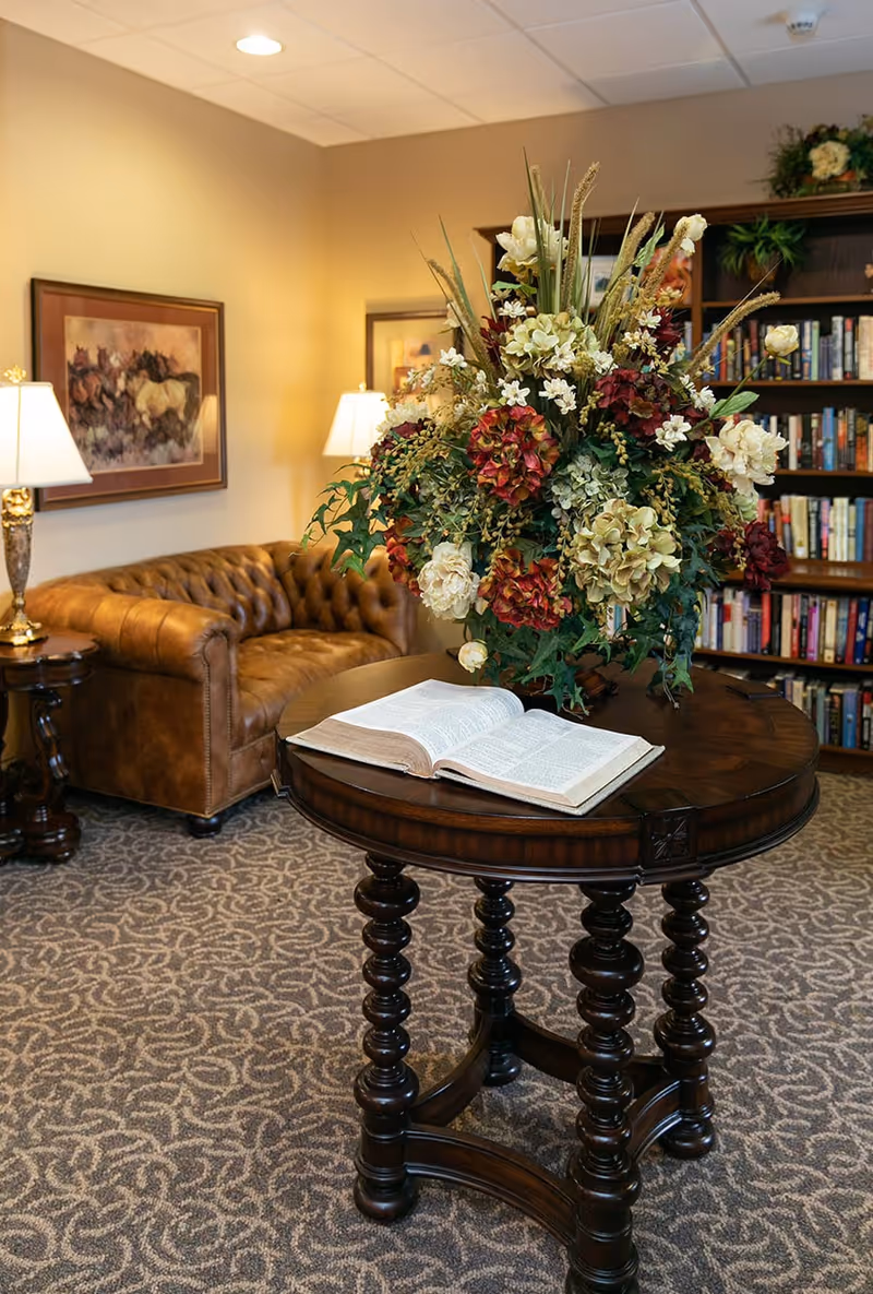 A cozy senior living common area with a brown leather tufted sofa, two table lamps, framed artwork on the wall, a round wooden table with a large floral arrangement and an open book, and a bookshelf filled with books in the background.