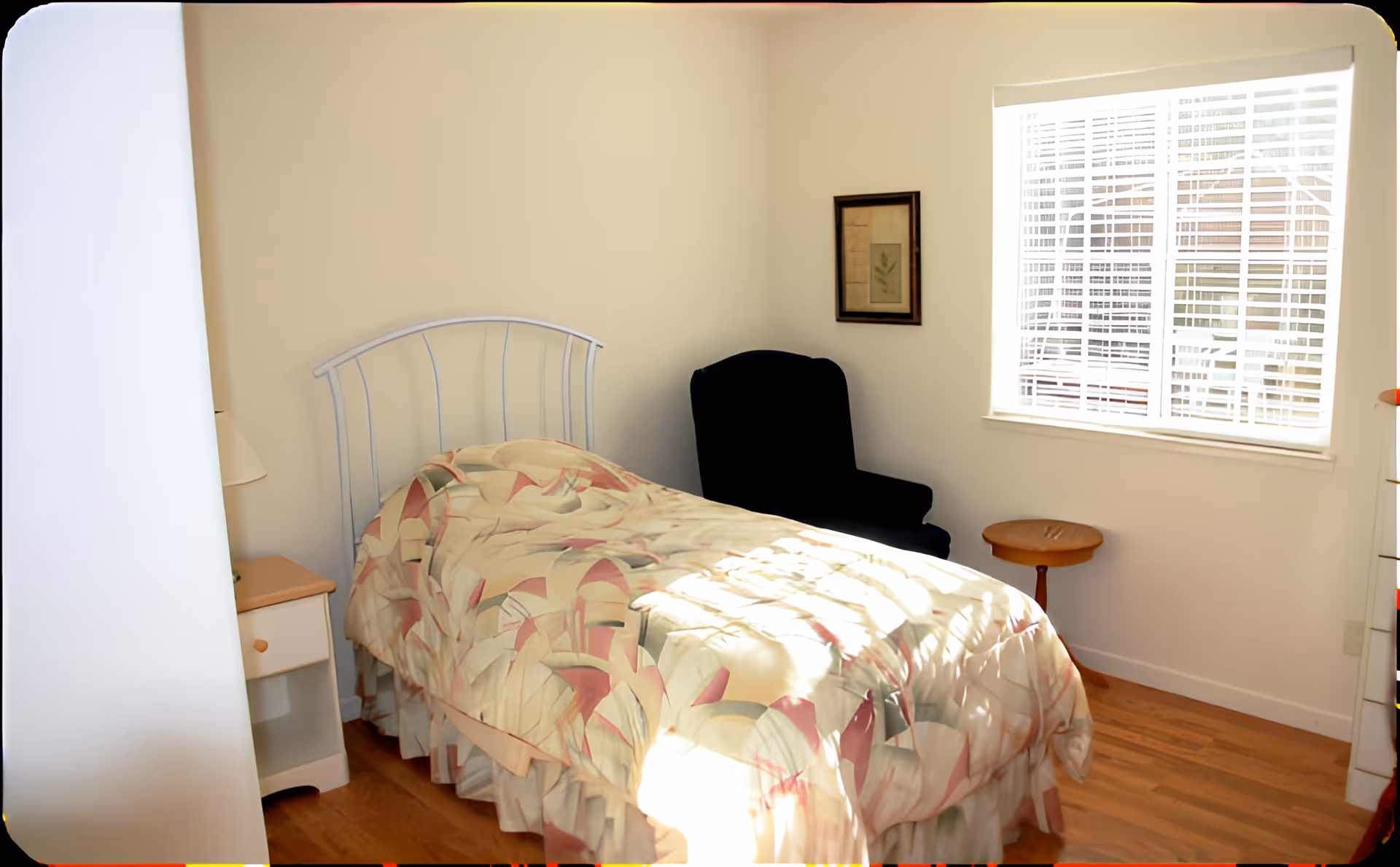 Sunlit single bedroom with a bed covered in a patterned bedspread, a black armchair, nightstand and a window with blinds.